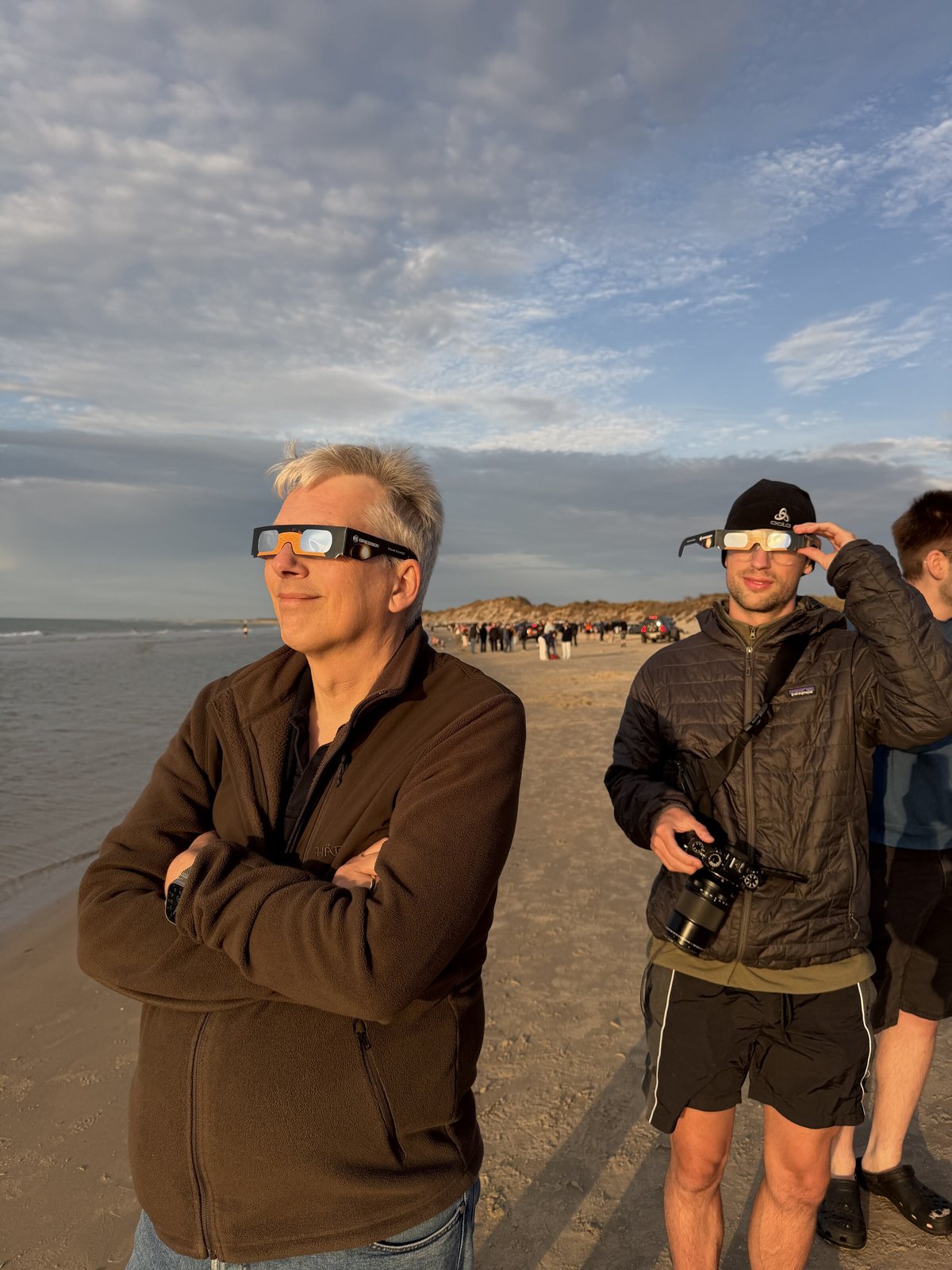 Strand von Tversted am Abend des 12. August 2026: zwei Zuschauer mit Sonnenfinsternisbrillen, im Hintergrund weitere Menschen und geparkte Autos an den D&uuml;nen